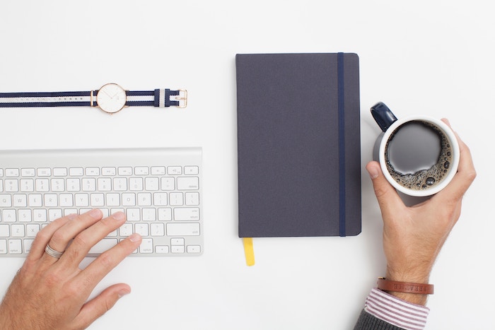 A man's hand holding a cup of coffee on an organised company workspace with a notebook and a keyboard