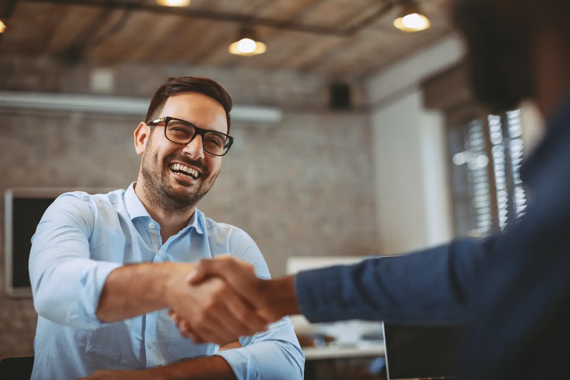 Two coworkers shake hands in a modern office, both smiling.